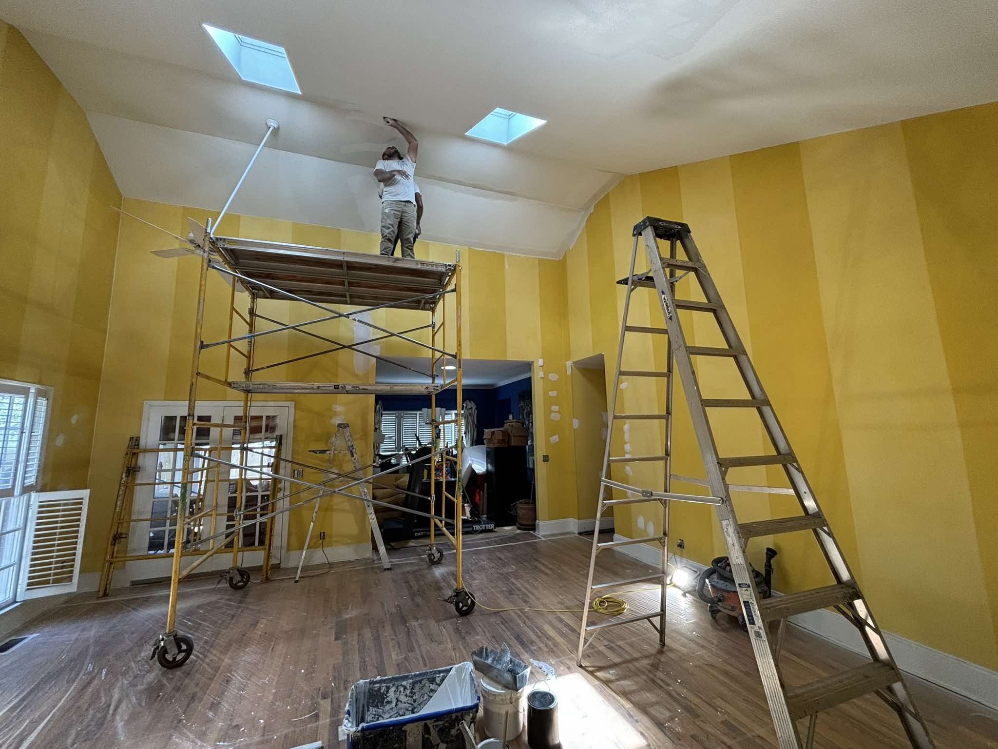 High-ceiling room during painting with yellow walls, rolling scaffold, ladders, and protected wood floors